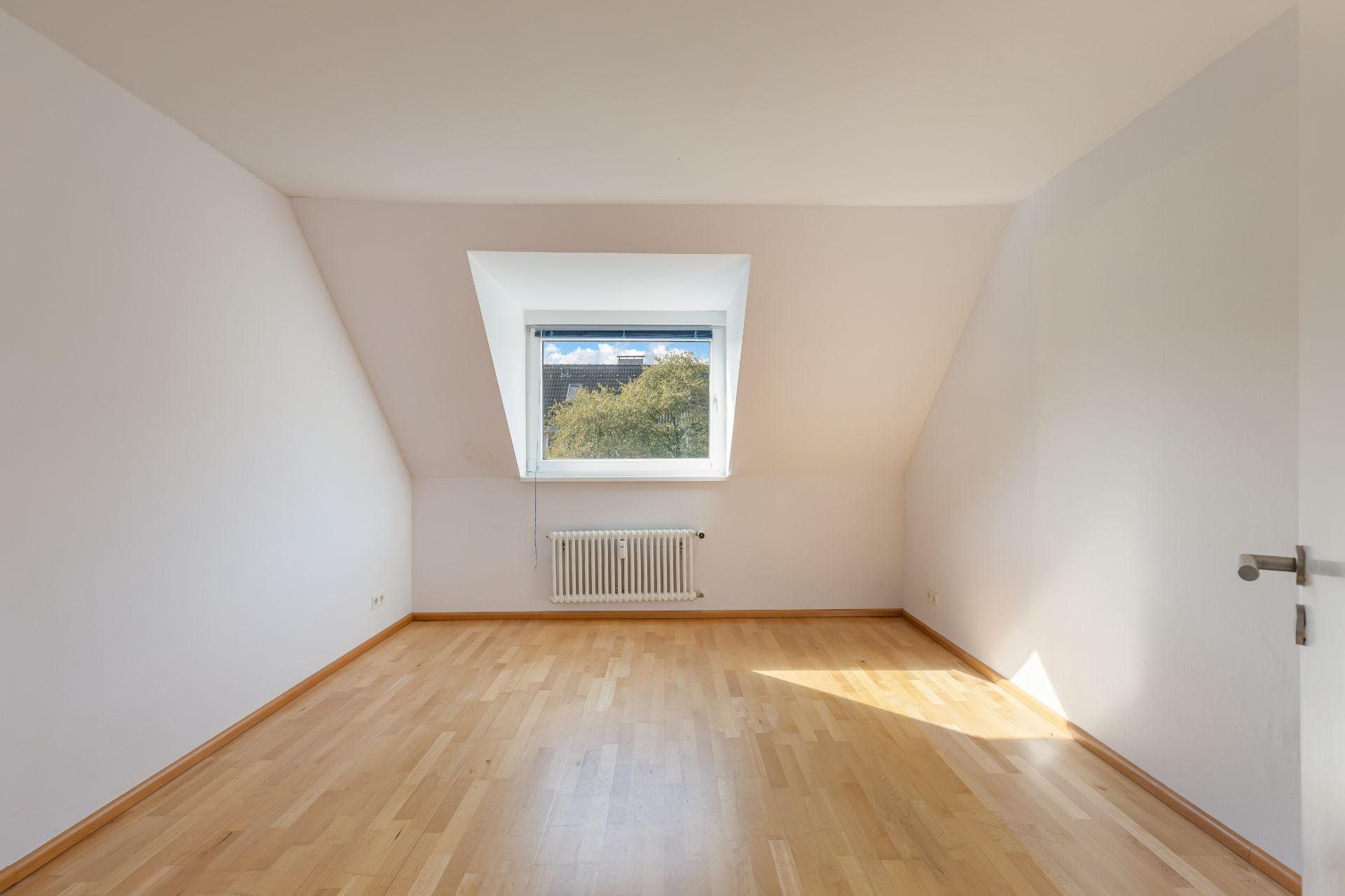 Leeres Zimmer mit schrägen Wänden, Holzfußboden und Fenster mit Blick ins Grüne.