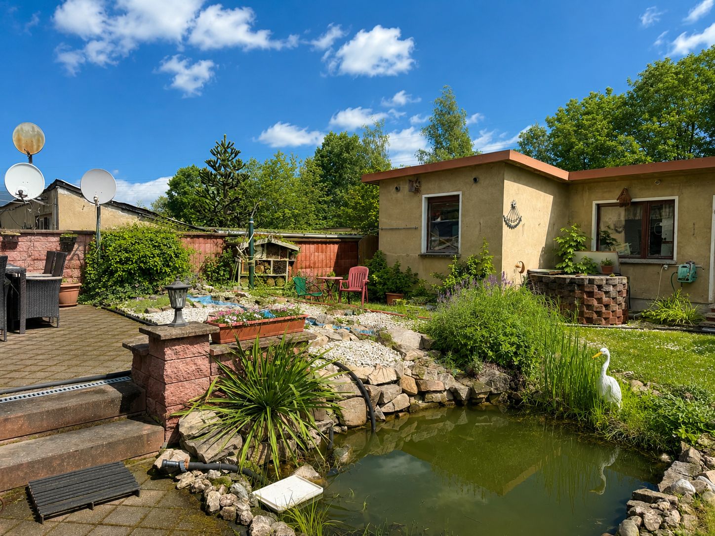 Garten mit kleinem Teich, Steinen, Gartenmöbeln und einem einstöckigen Haus im Hintergrund.