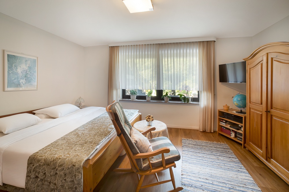 Helles Schlafzimmer mit Doppelbett, Schaukelstuhl, Teppich, Holzschrank und großem Fenster mit Vorhängen.