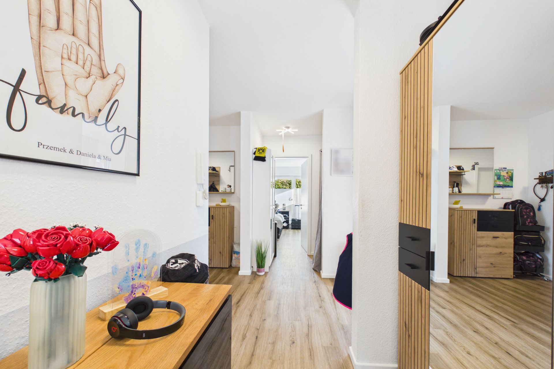 Heller Flur mit Holzboden, Kommode, Spiegel, Blumen und Blick in ein weiteres Zimmer.