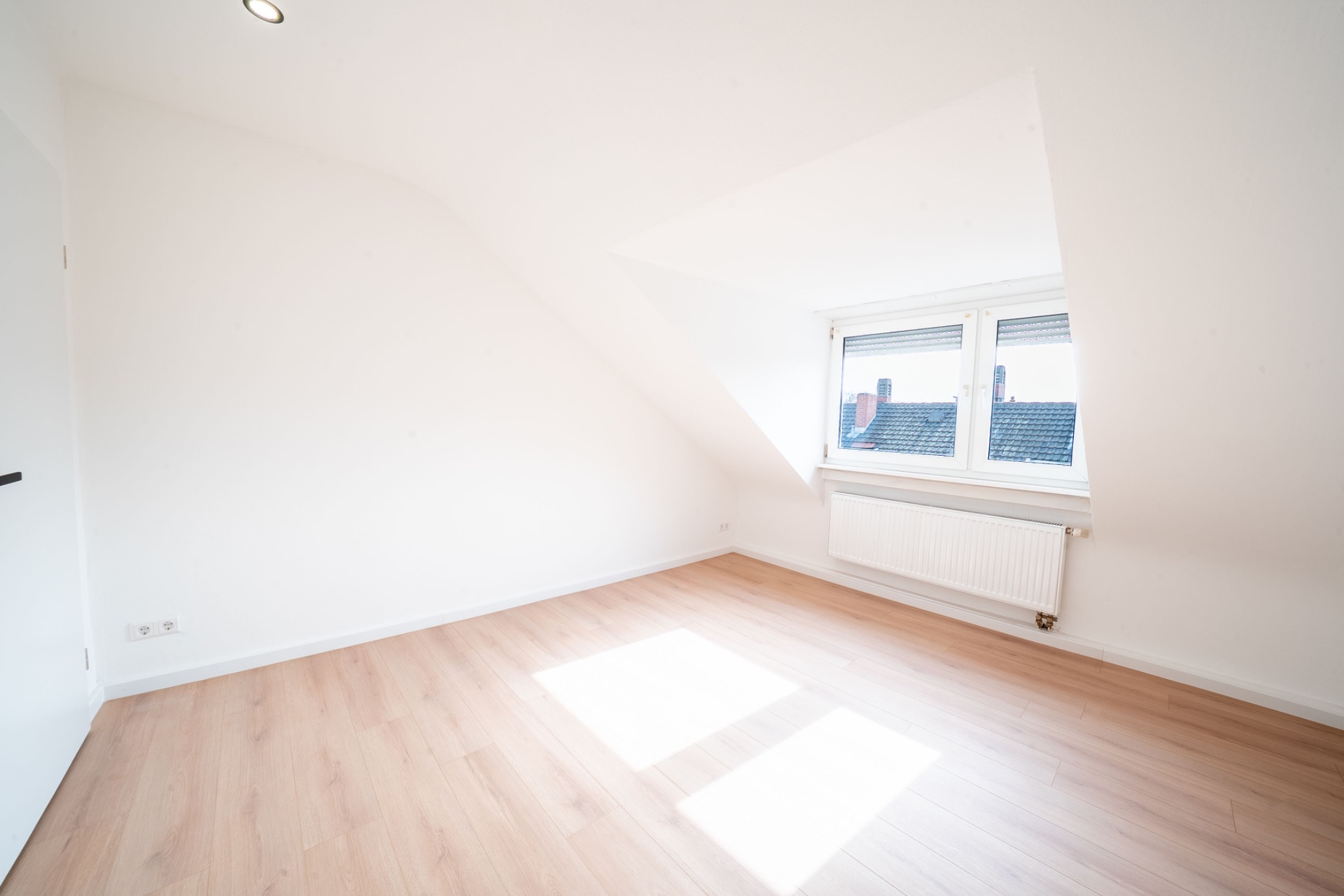 Helles, leeres Zimmer mit schräger Decke, Holzfußboden und großem Fenster mit Heizung darunter.