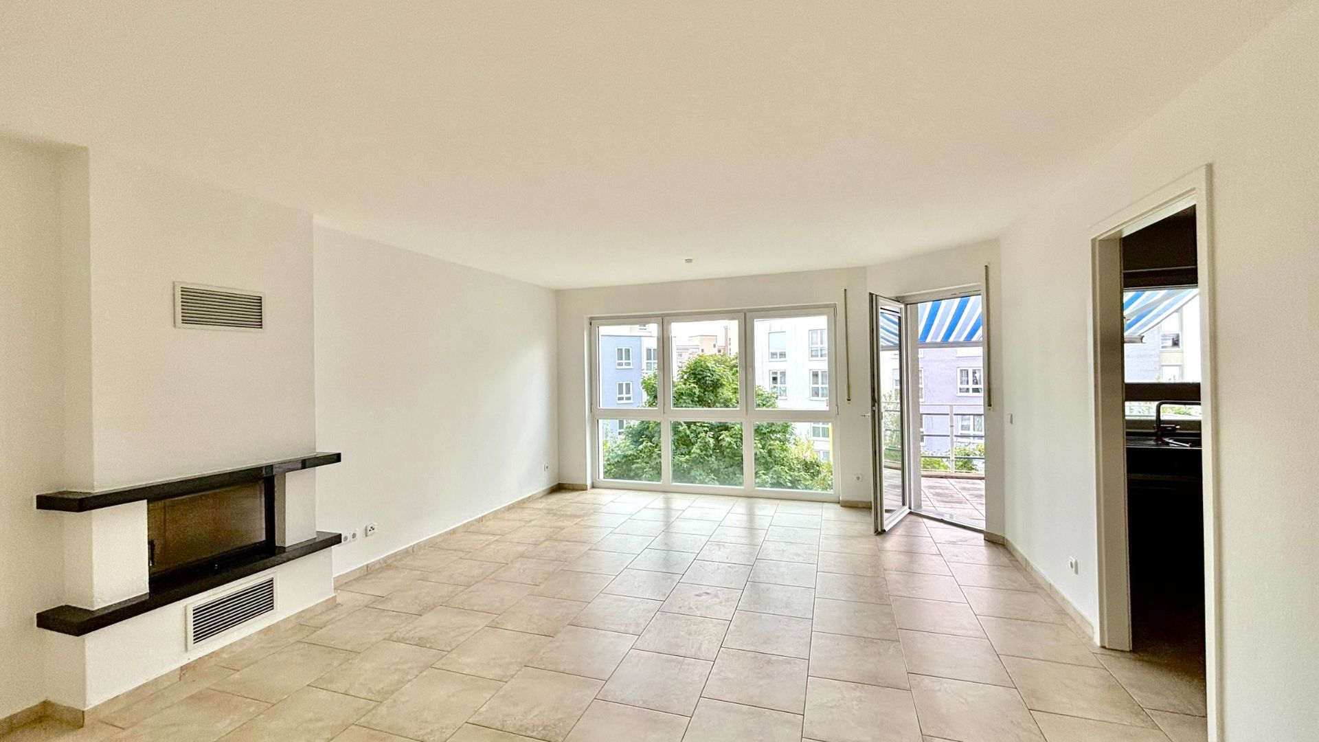 Helles Wohnzimmer mit Kamin, großen Fenstern, Fliesenboden und Zugang zum Balkon.