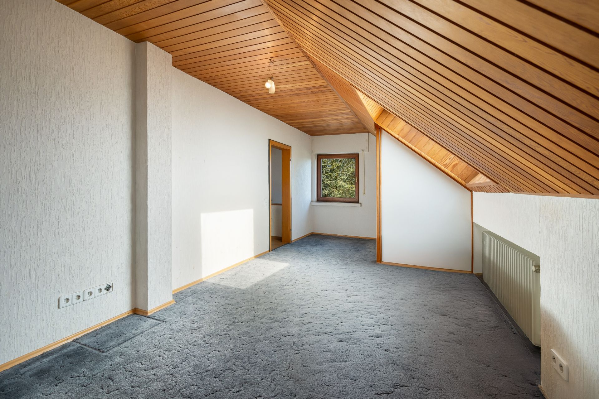 Dachzimmer mit Holzdecke, schräger Wand, Teppichboden, Fenster und Heizkörper.