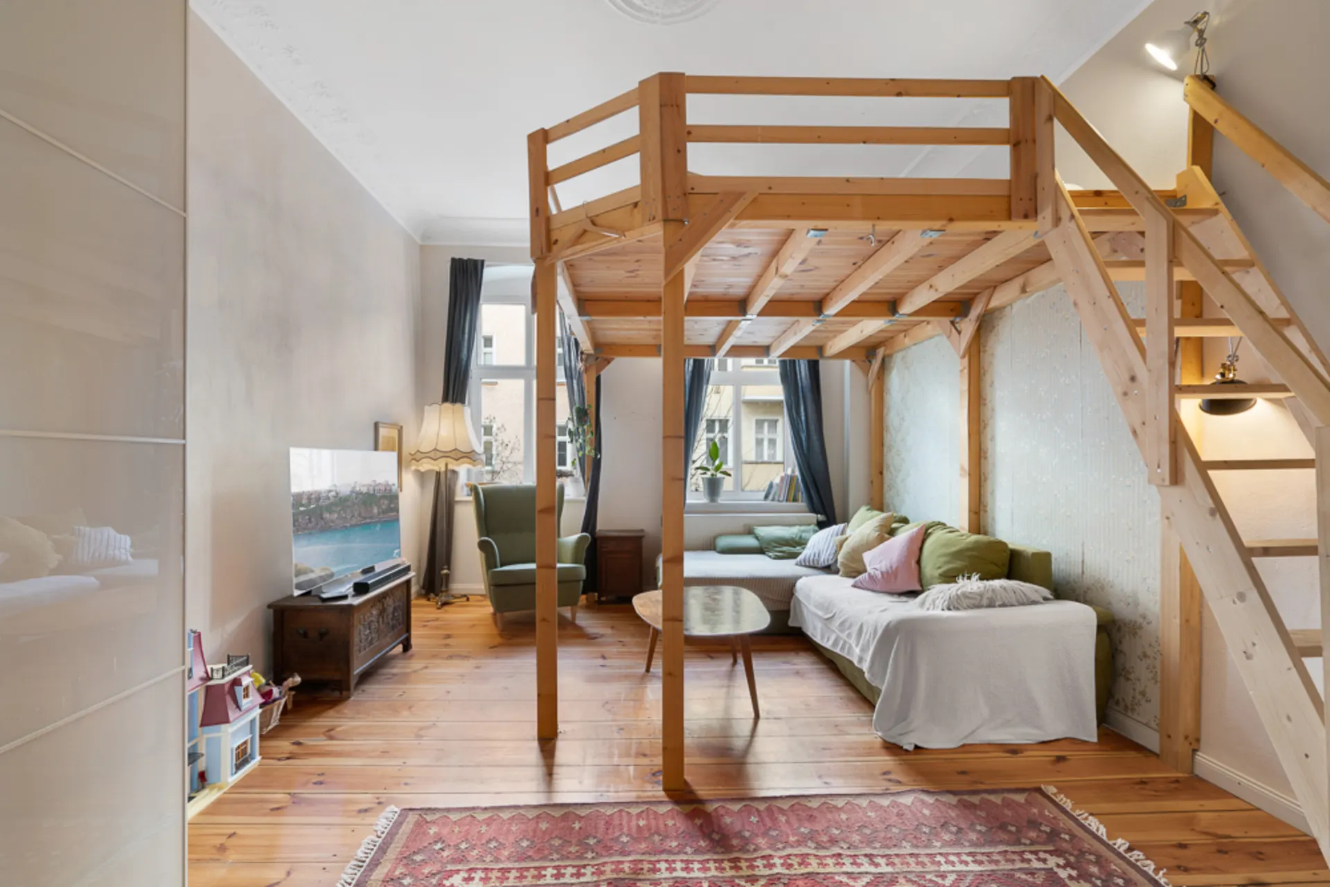 Wohnzimmer mit Hochbett, Sofa, Sessel, TV, Holzboden und großem Fenster.