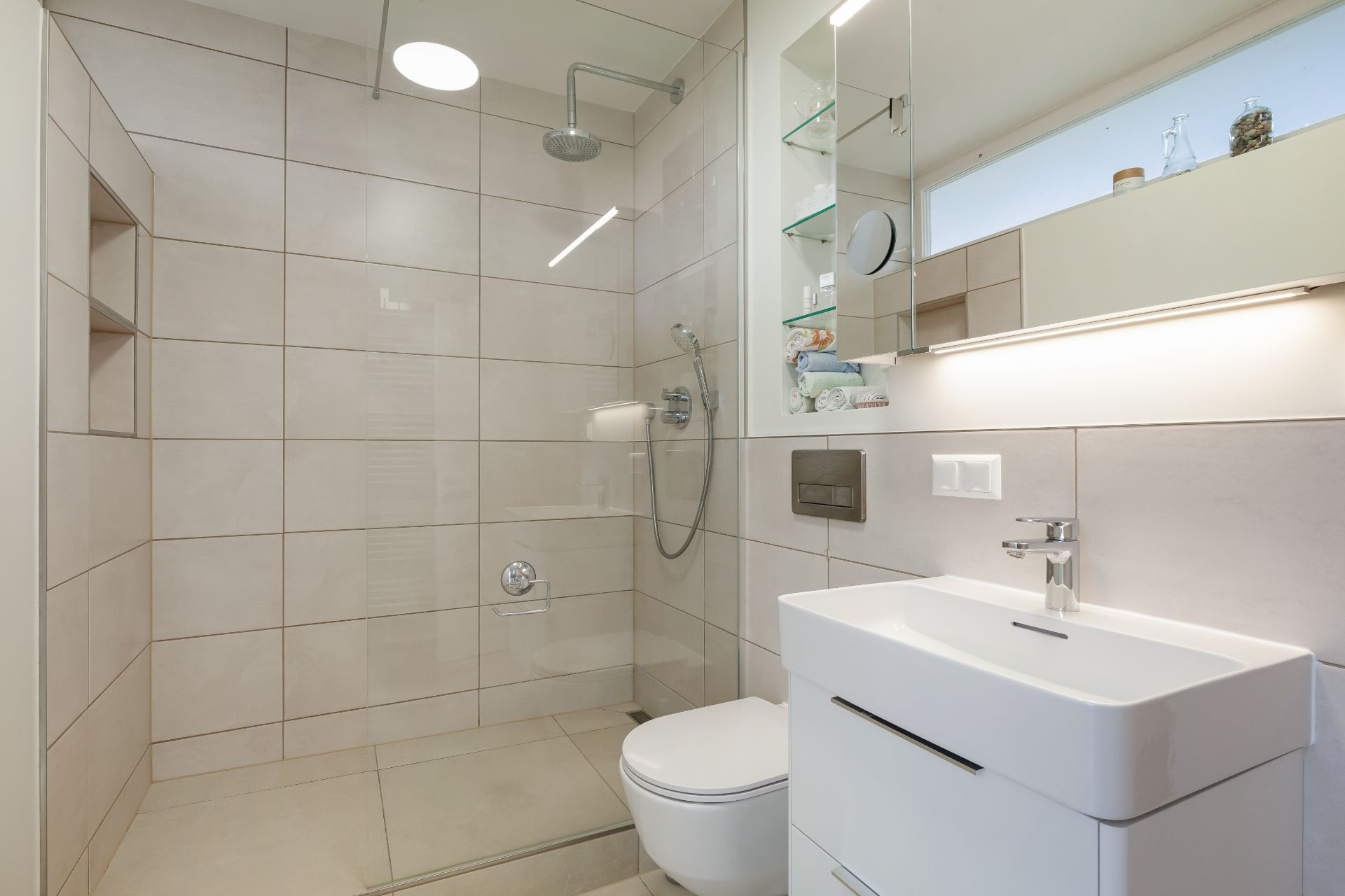 Modernes Badezimmer mit ebenerdiger Dusche, WC, Waschbecken und beleuchtetem Spiegel.
