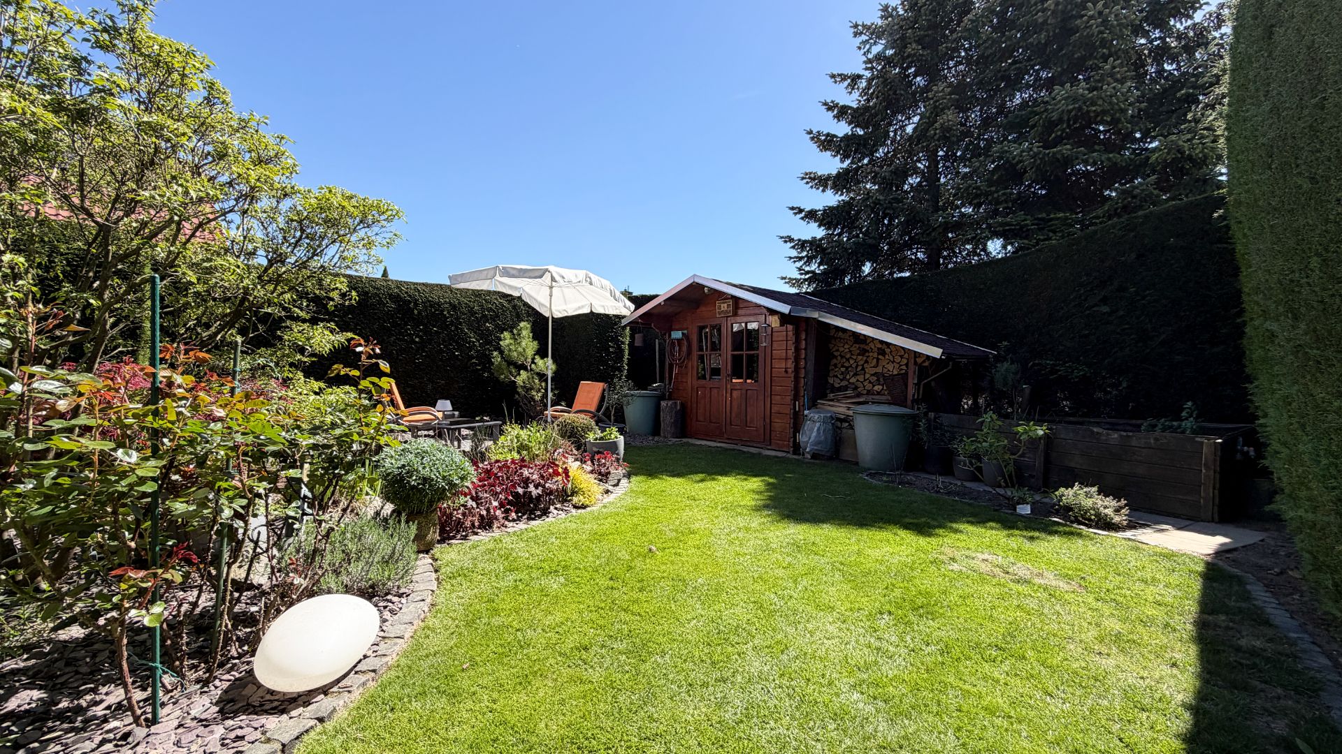 Grüner Garten mit Holzhaus, Blumenbeet und Gartenmöbeln unter einem Sonnenschirm.