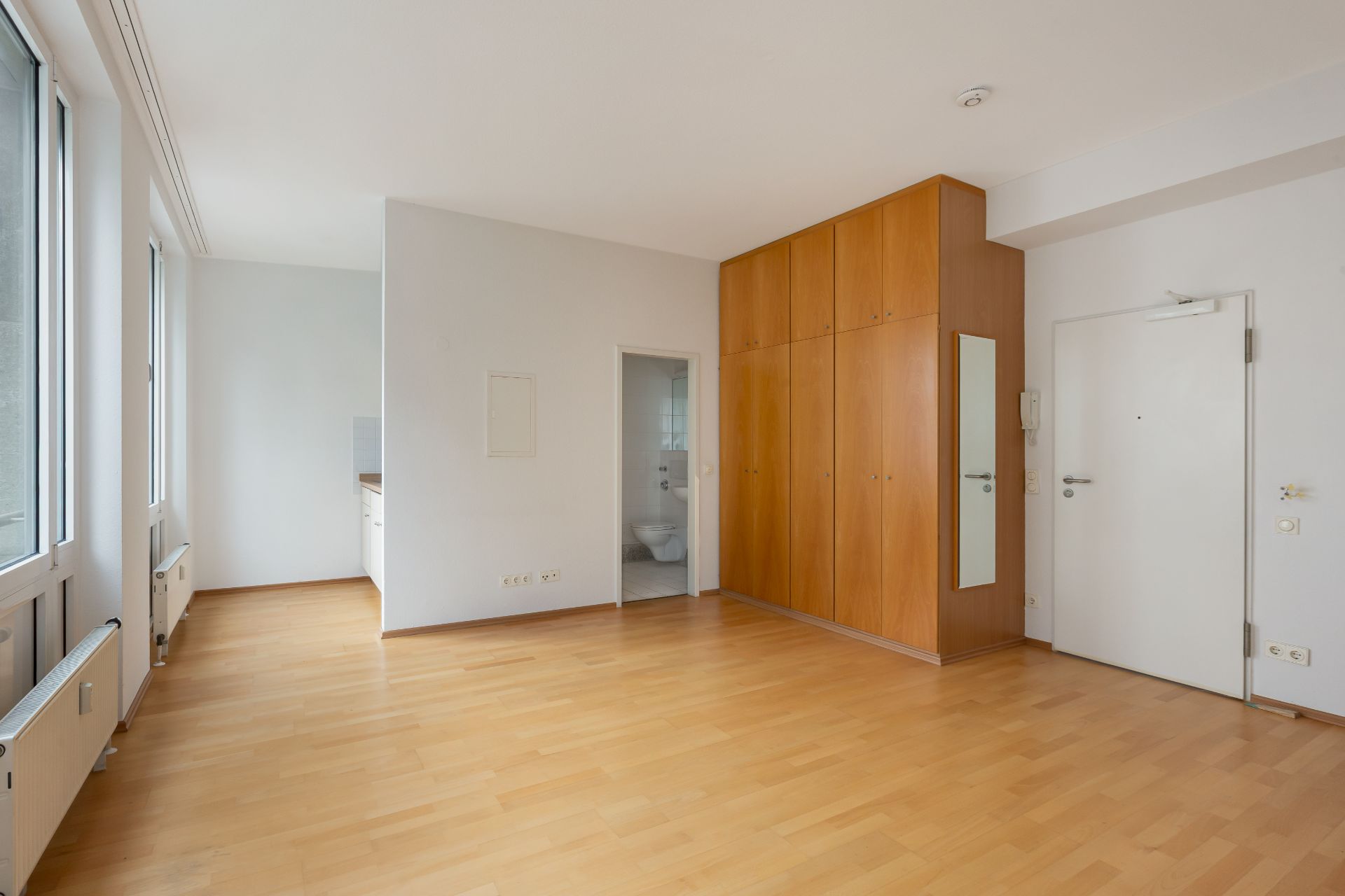Heller Raum mit großem Einbauschrank, Spiegel, Holzboden und Zugang zur Küche.