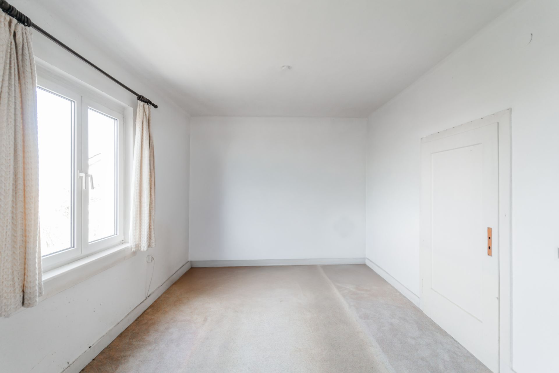 Leeres, helles Zimmer mit Teppichboden, Fenster mit Vorhängen und einer weißen Tür.