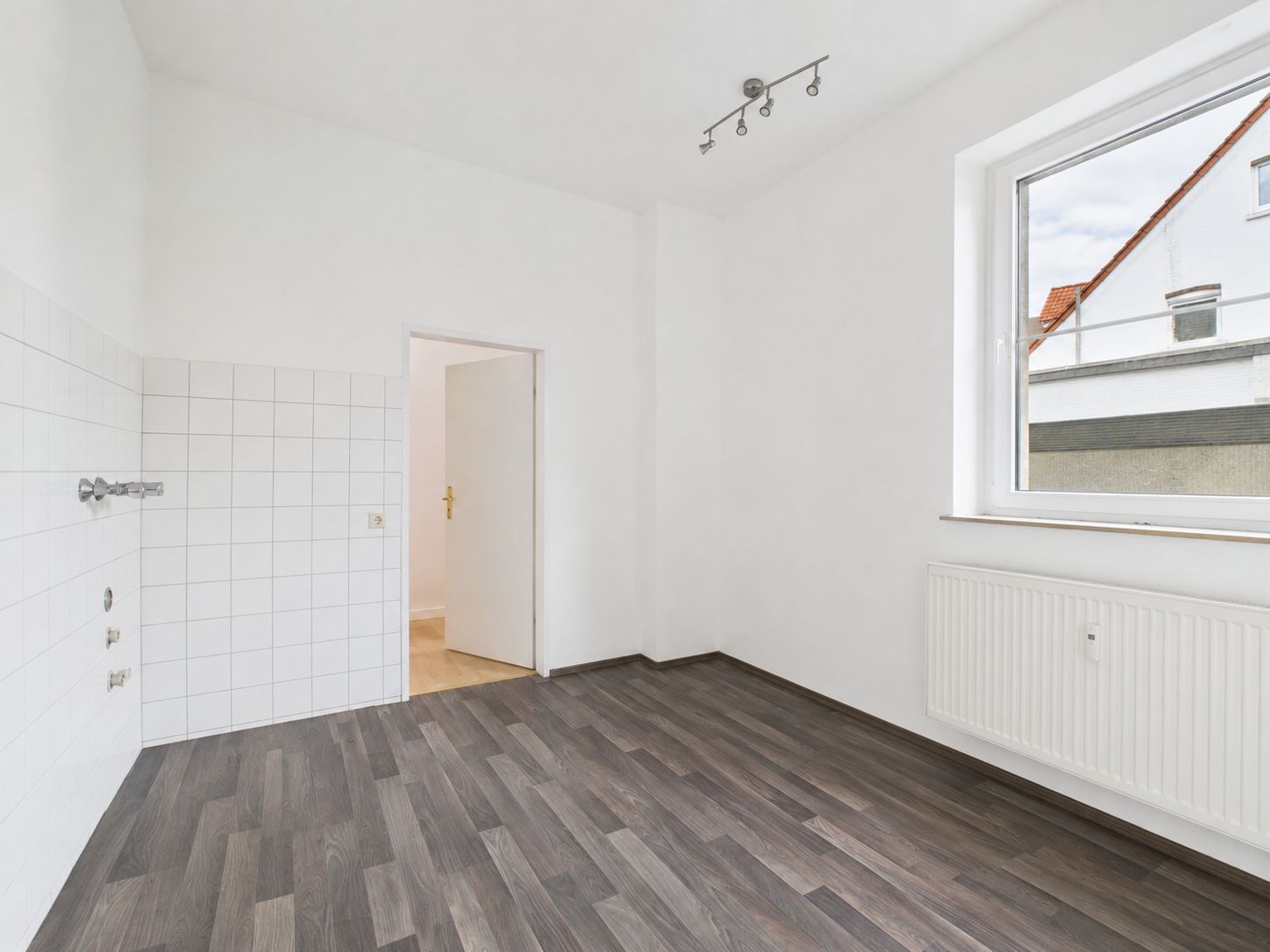 Leerer Raum mit dunklem Boden, weißer Wand, Fenster, Heizung und Tür.