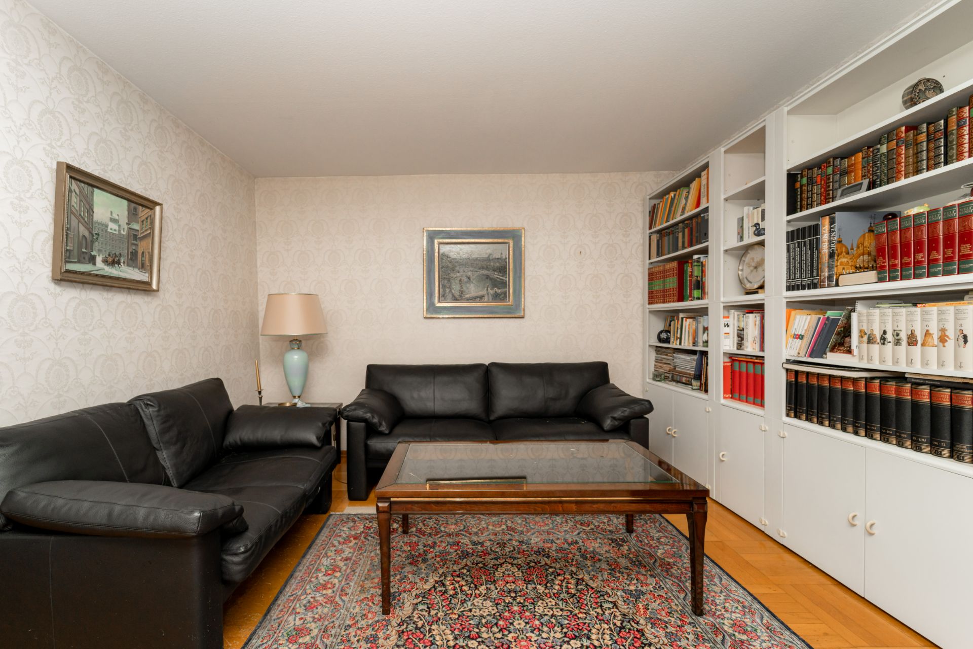 Wohnzimmer mit zwei schwarzen Ledersofas, Glas-Couchtisch, Teppich und Bücherregal an der Wand.