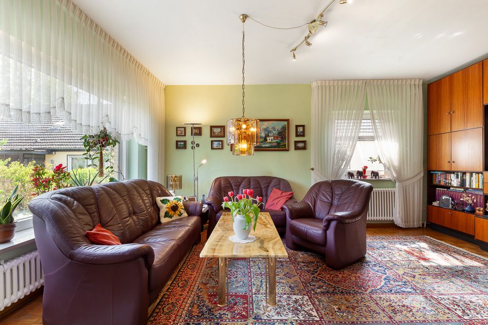 Wohnzimmer mit braunen Ledersofas, Holztisch, Teppich und großen Fenstern mit Vorhängen.