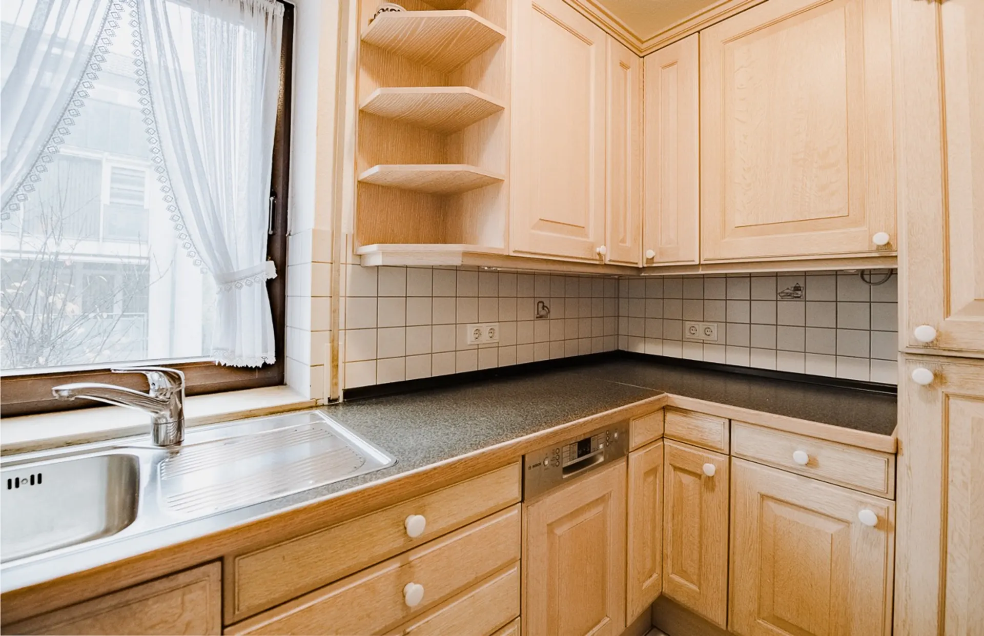 Helle Einbauküche mit Holzfronten, Eckspüle, Geschirrspüler und Fenster mit Spitzevorhang.