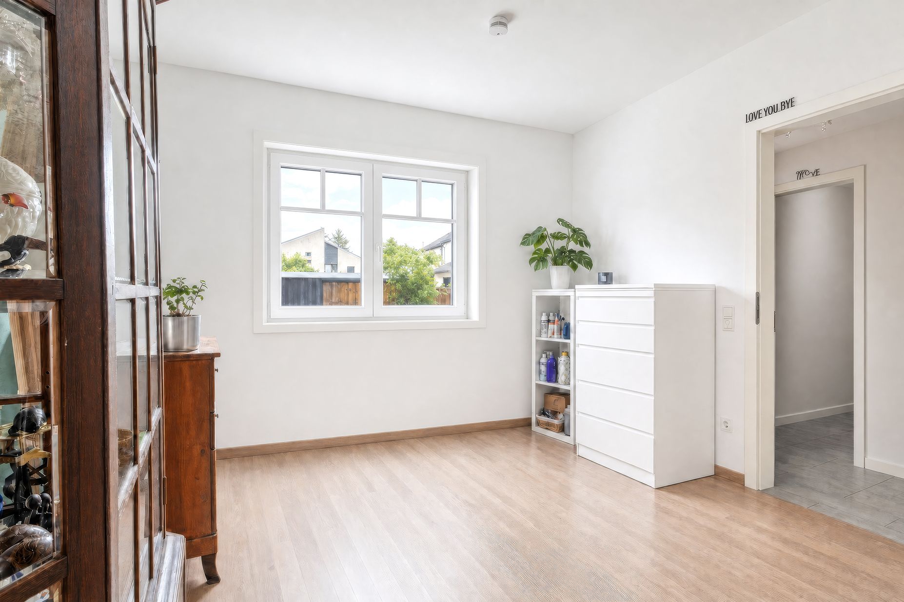 Helles Zimmer mit Holzfußboden, Fenster, Kommode, Regal und Pflanze.