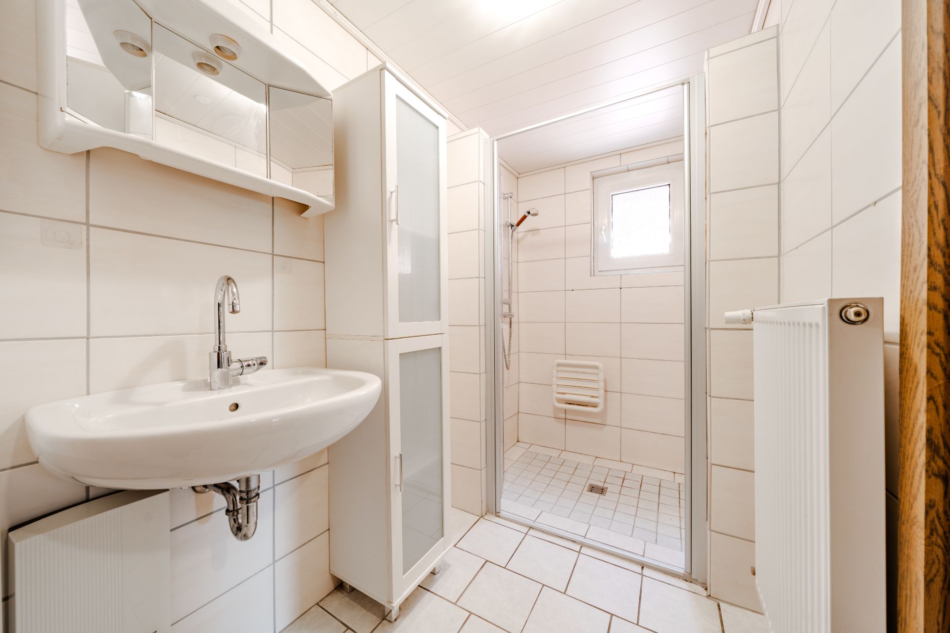 Helles Badezimmer mit Waschbecken, Spiegel, Duschkabine, Fenster und Heizkörper.