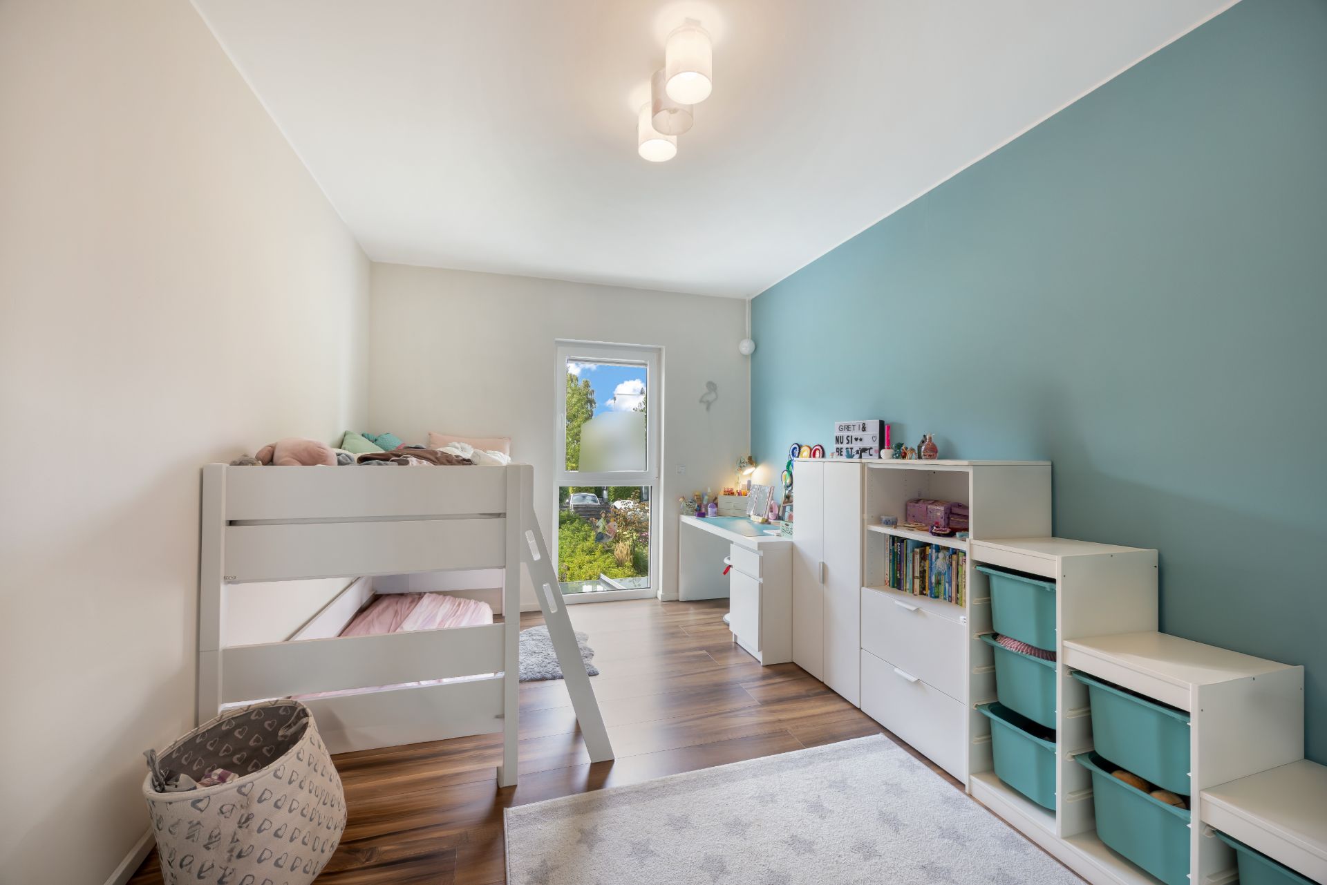 Helles Kinderzimmer mit Hochbett, Schreibtisch, Regalen und Spielzeug, Holzboden und blau gestrichener Wand.