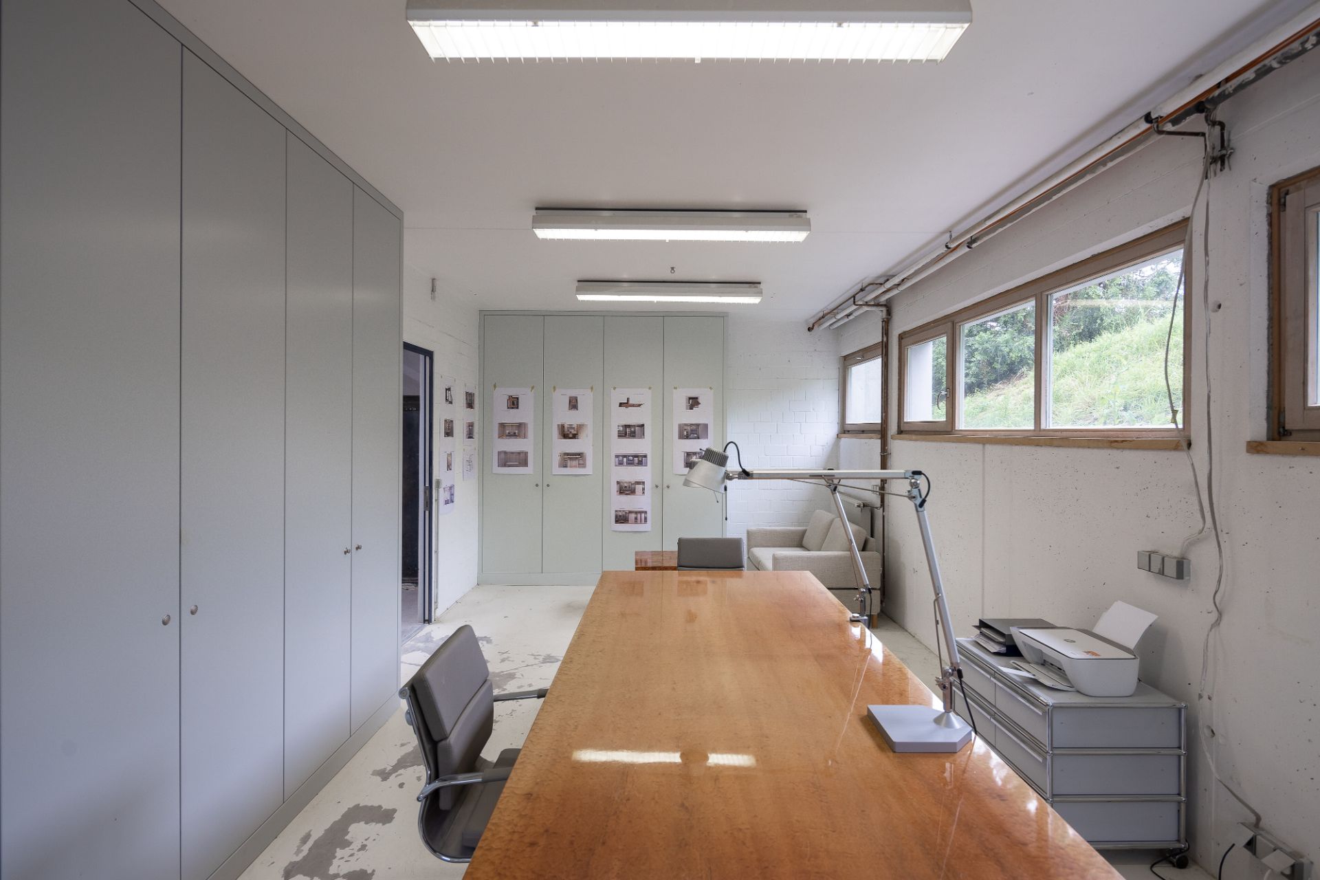 Helles Arbeitszimmer mit großem Holztisch, Stuhl, Schreibtischlampe und Fenstern an der rechten Wand.