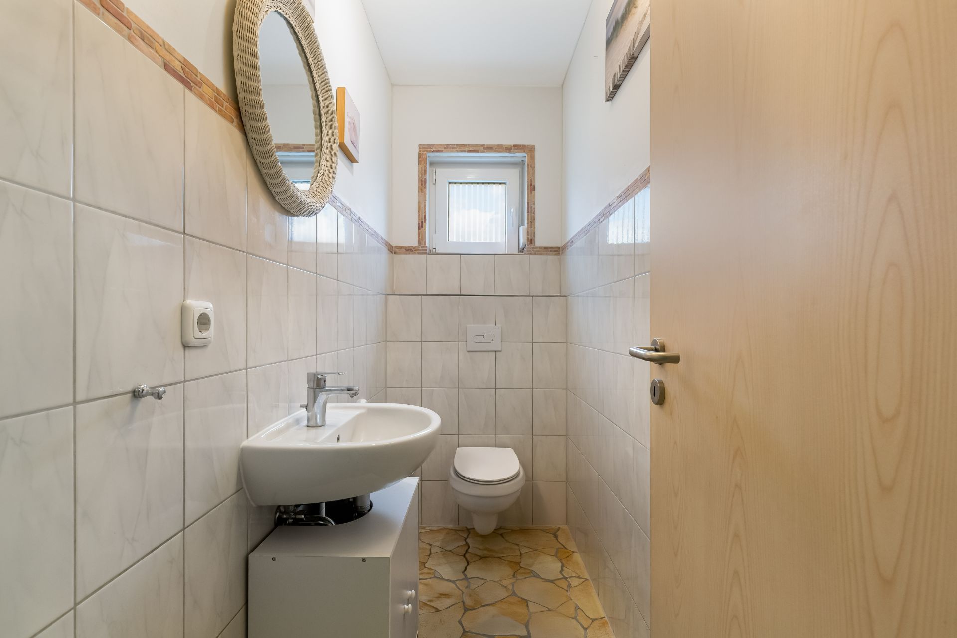 Kleines Badezimmer mit Wandtoilette, Waschbecken und rundem Spiegel, gefliest.