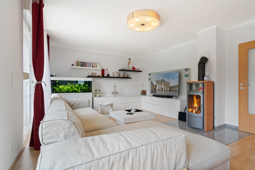 Helles Wohnzimmer mit weißem Sofa, Aquarium, Kaminofen und Fernseher an der Wand.