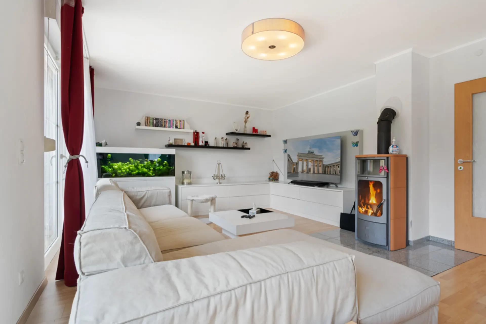 Helles Wohnzimmer mit weißem Sofa, Aquarium, Kaminofen und Fernseher an der Wand.