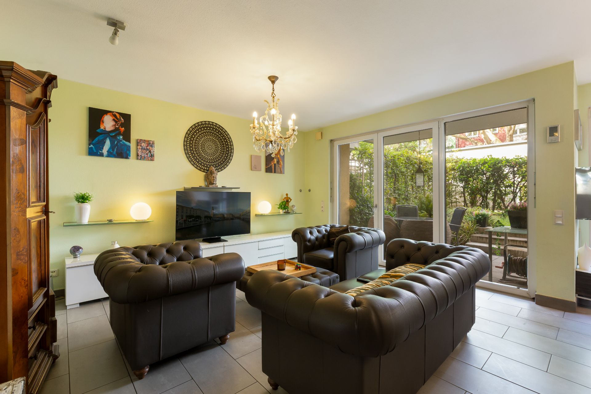 Wohnzimmer mit drei Ledersofas, Fernseher, Kronleuchter und Zugang zur Terrasse mit Garten.