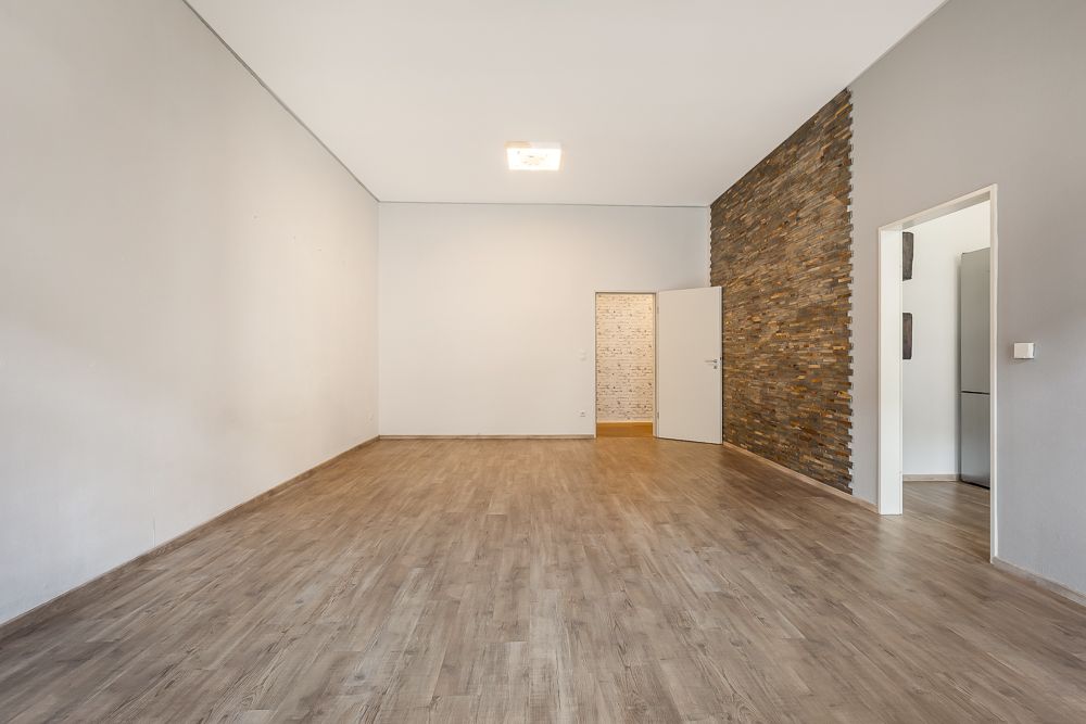 Leeres Zimmer mit Holzfußboden und Steinwand, offene Tür rechts zur angrenzenden Küche.
