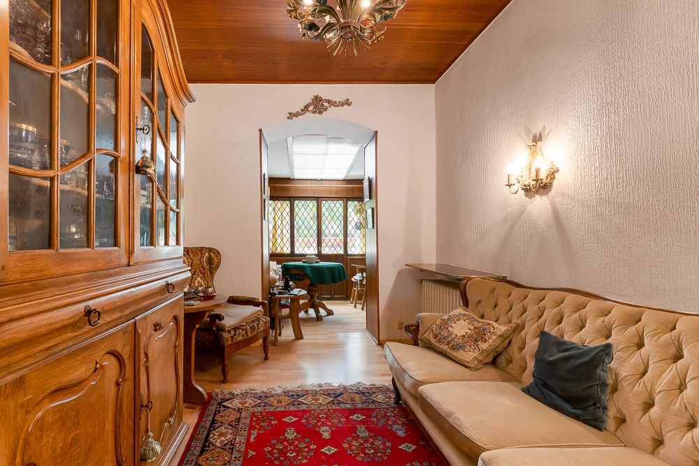 Wohnzimmer mit Holzmöbeln, cremefarbenem Sofa, Teppich und Blick auf Essbereich mit bunten Fenstern.