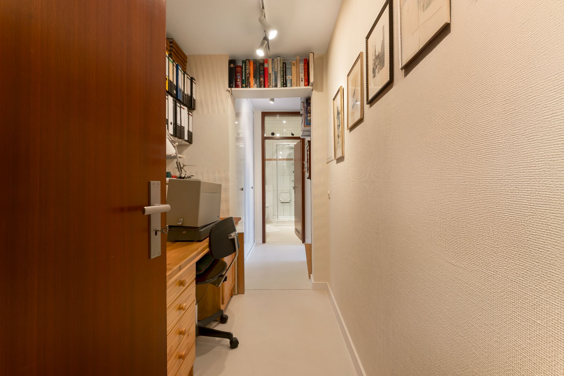 Schmaler Flur mit Schreibtisch, Regalen und Bildern an der Wand, Blick ins Badezimmer.