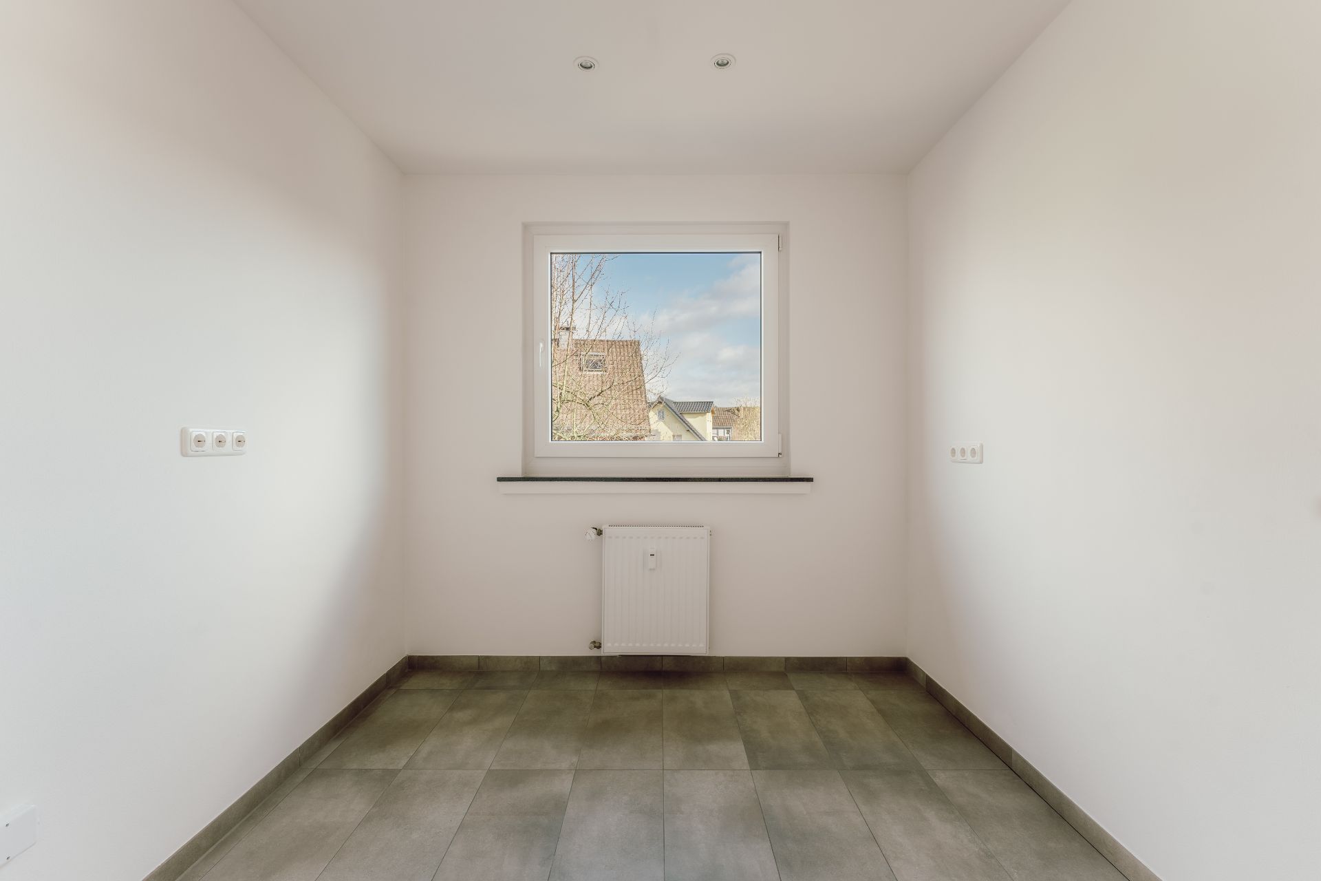 Leerer, heller Raum mit Fliesenboden, weißer Wand, Fenster und Heizkörper.