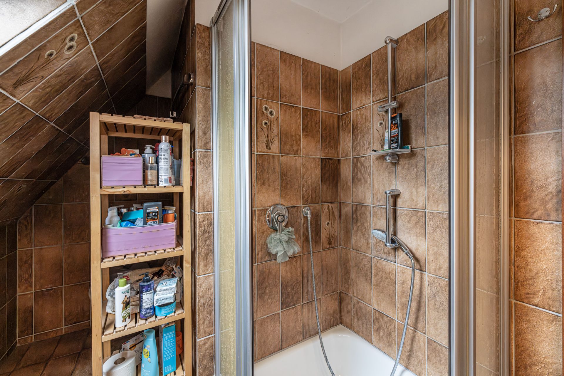 Badezimmer mit braunen Fliesen, Dusche und Regal mit Pflegeprodukten.