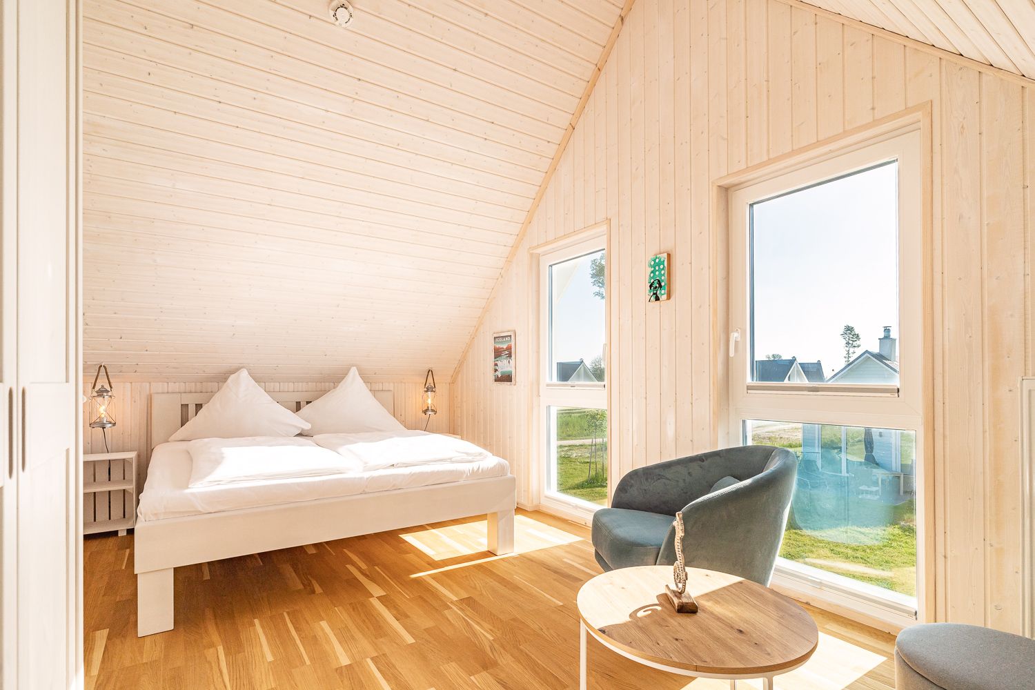 Helles Schlafzimmer mit Doppelbett, zwei Fenstern, Sessel und Holztisch auf Holzfußboden.