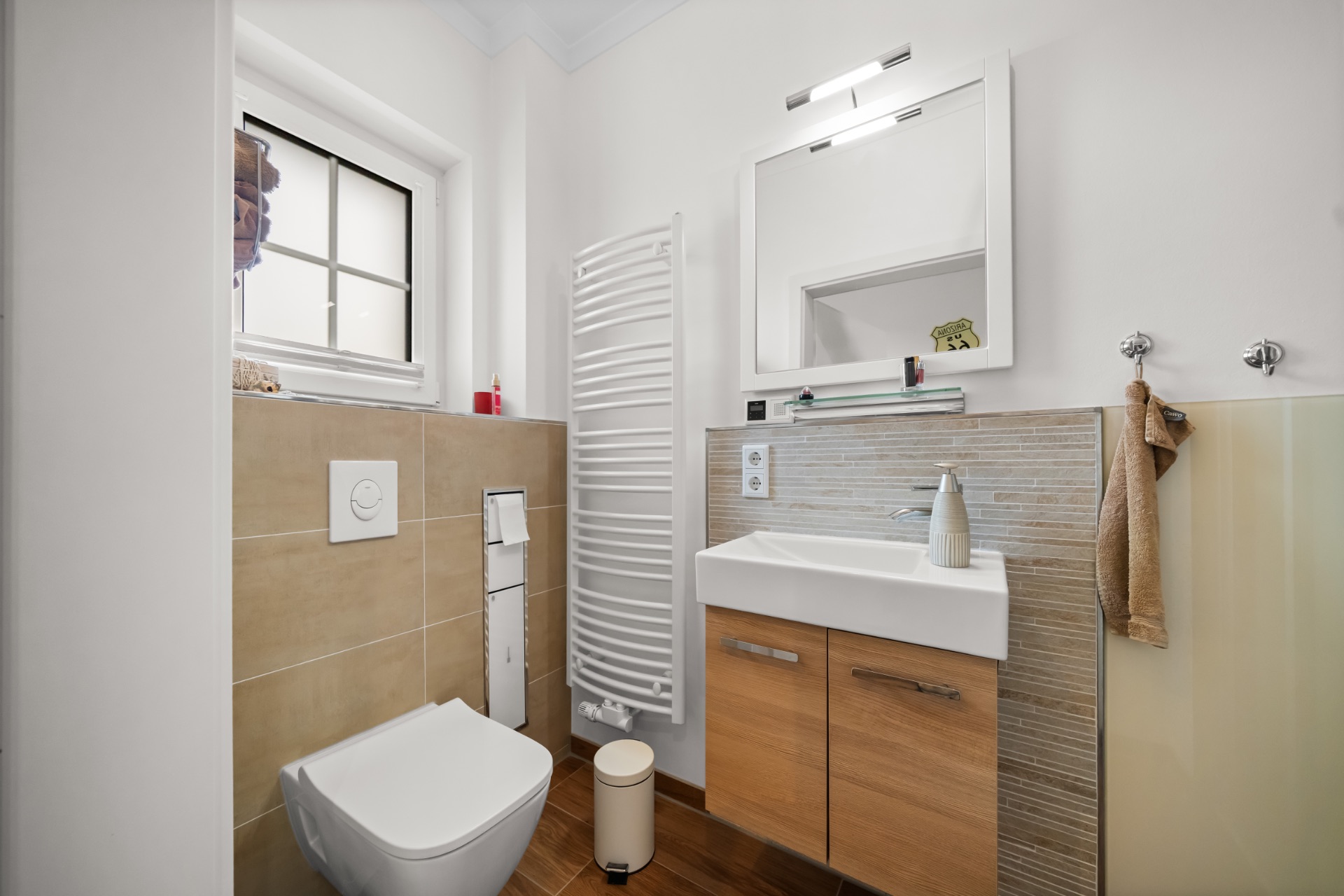 Modernes Badezimmer mit Wandtoilette, Waschbecken, Spiegel und Handtuchheizung.