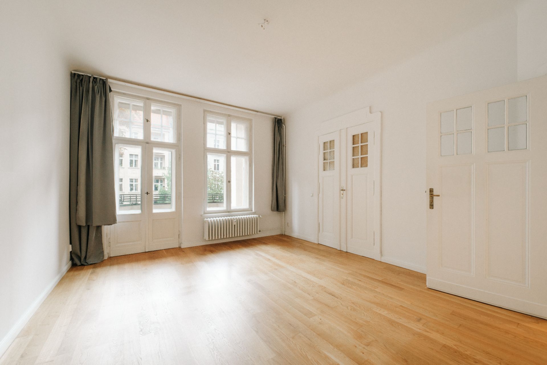 Helles, leeres Zimmer mit Holzboden, großen Fenstern, Vorhängen und weißen Türen.