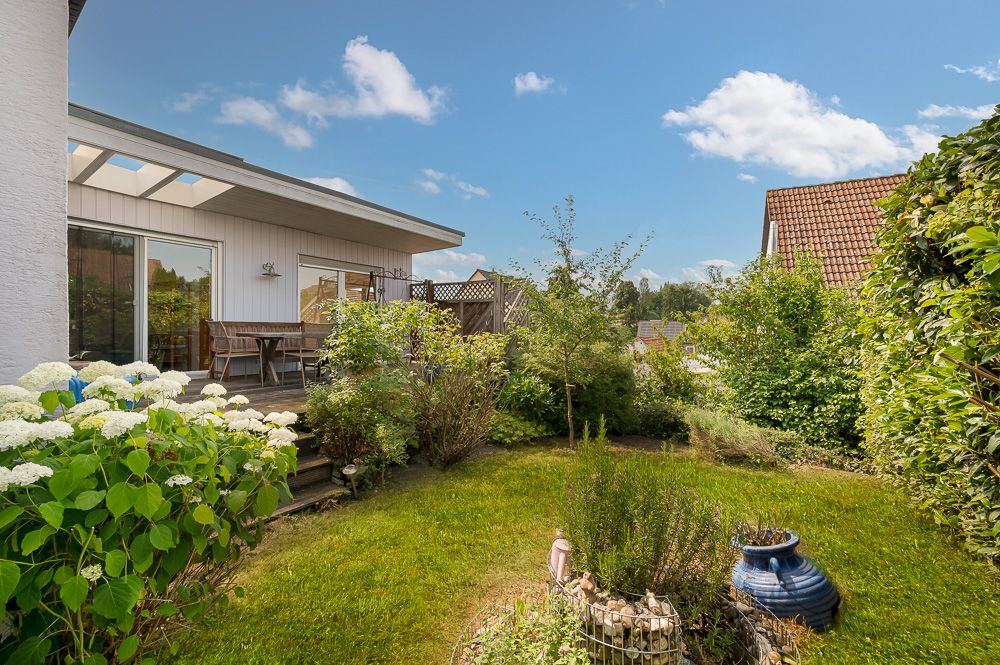 Grüner Garten mit Terrasse, Gartenmöbeln, Blüten und Sträuchern an einem Wohnhaus bei Sonnenschein.