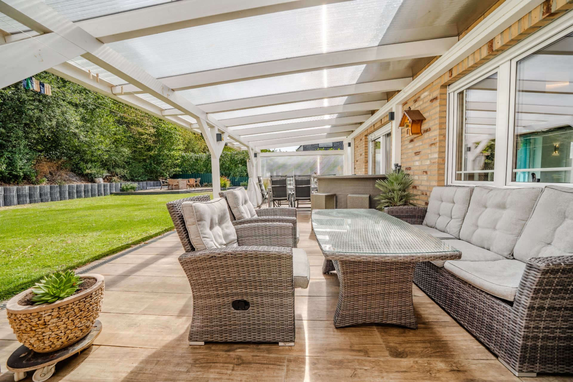 Überdachte Terrasse mit Sitzgruppe aus Rattanmöbeln, Tisch und Blick auf einen Garten.