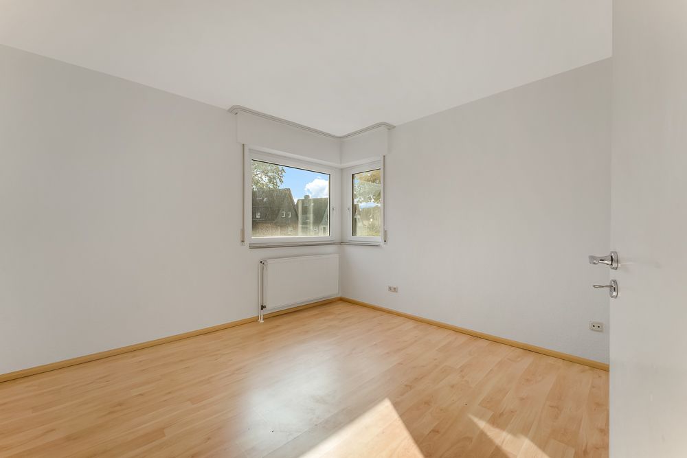 Helles, leeres Zimmer mit Laminatboden und zwei Fenstern in einer Ecke.