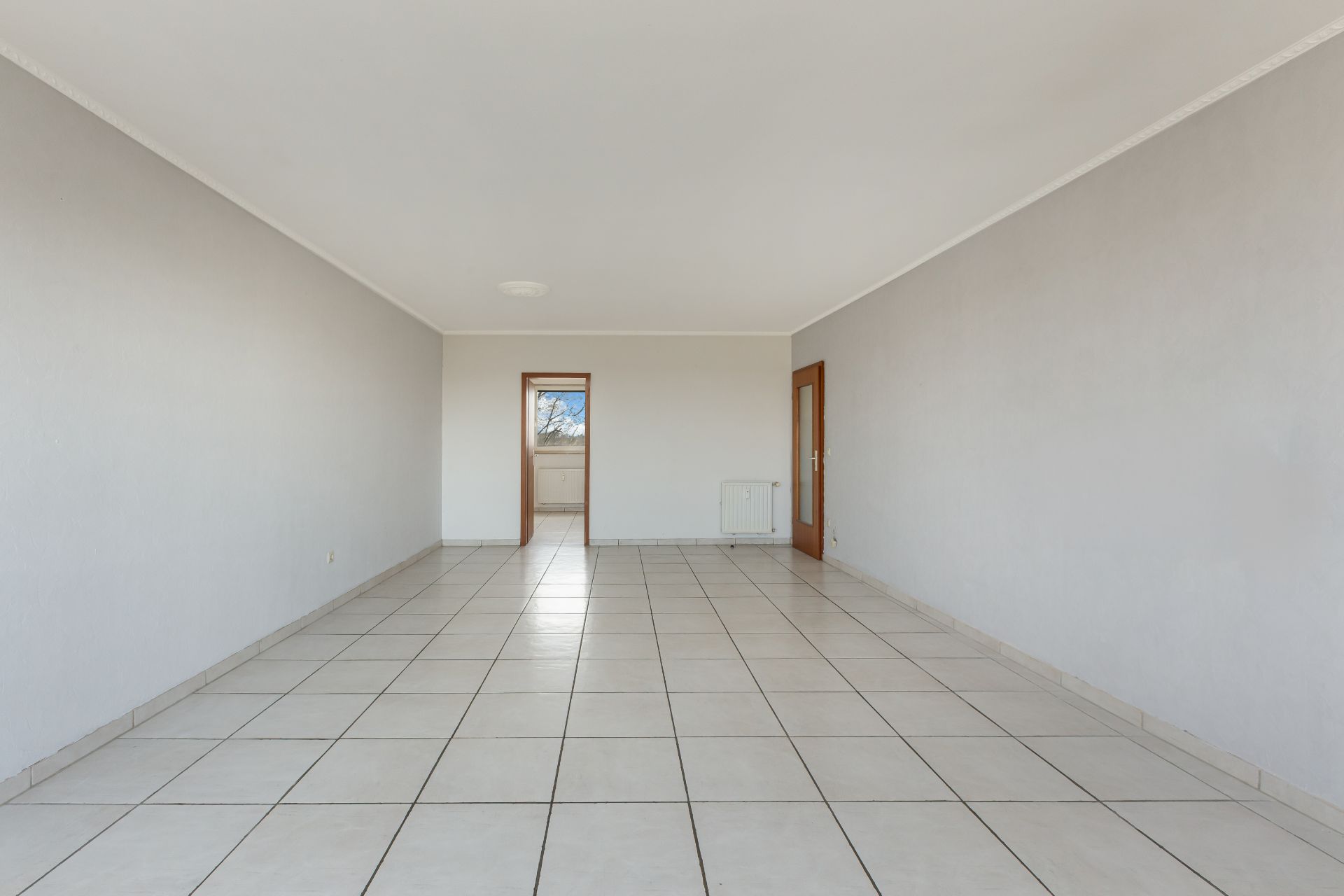 Leerer Raum mit weißen Fliesenboden, weißen Wänden und Tür mit Fenster im Hintergrund.