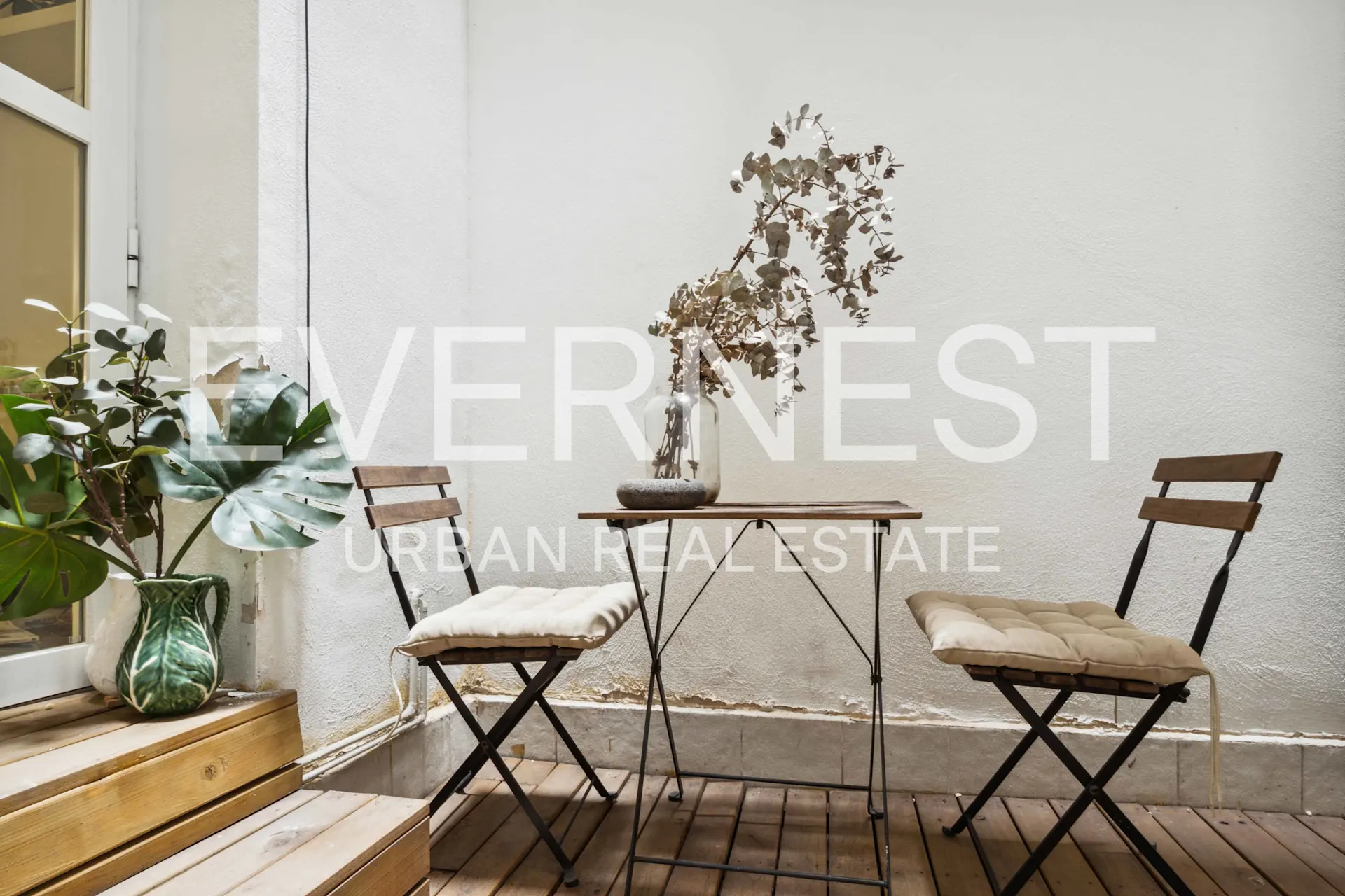 Kleiner Balkon mit Holzboden, zwei Stühlen, Tisch und Pflanzen an weißer Wand.