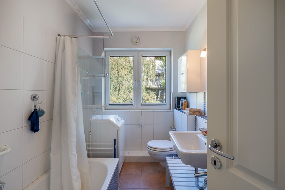 Helles Badezimmer mit Badewanne, Dusche, Waschbecken, WC und Fenster mit Blick nach draußen.