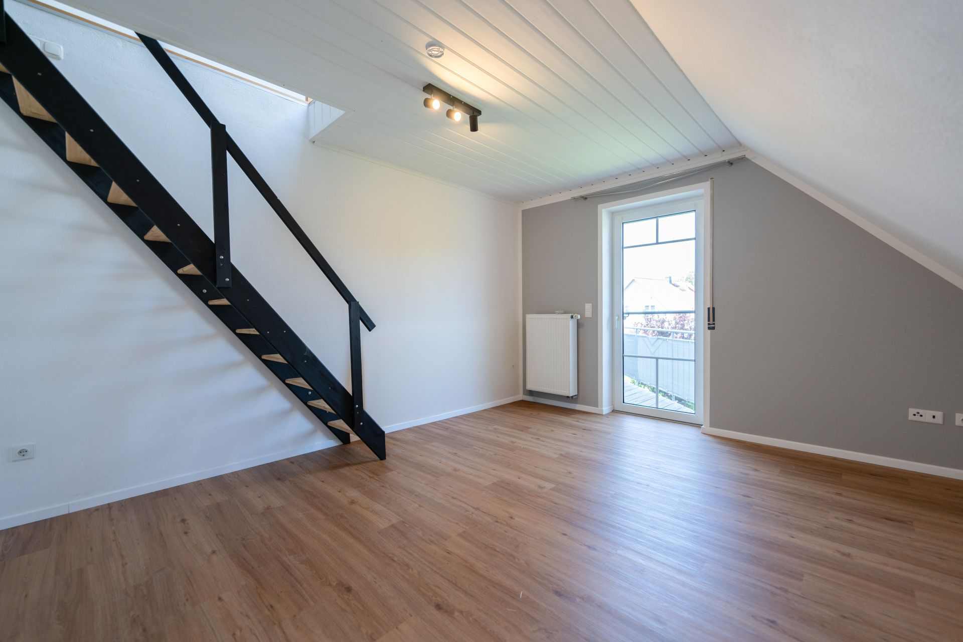 Helles Zimmer mit schräger Decke, Holzfußboden, schwarzer Treppe und Glastür zum Balkon.