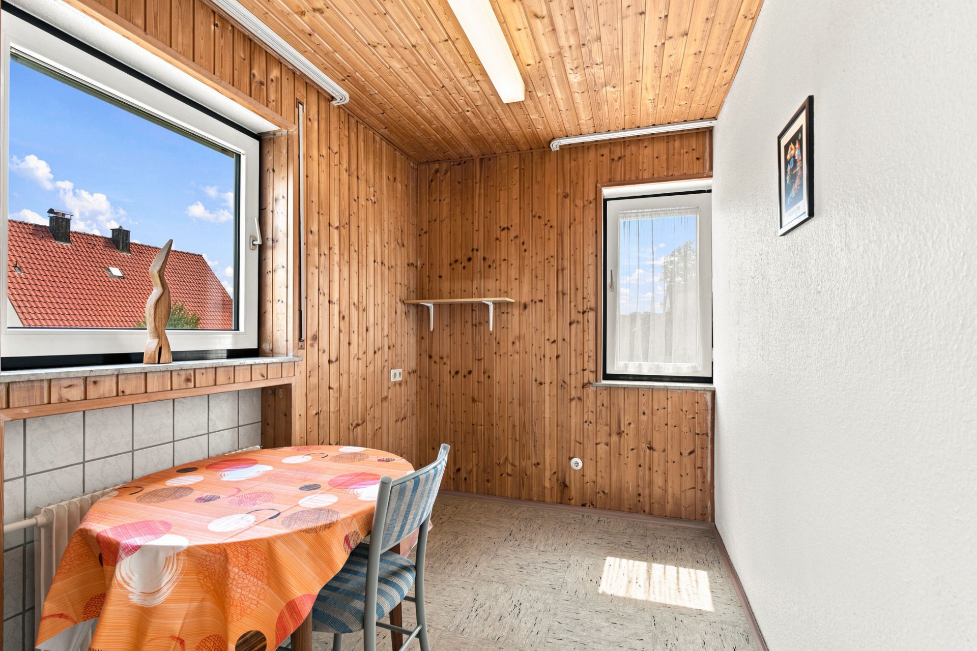 Kleiner Raum mit Holzwänden, Tisch mit orangener Tischdecke, zwei Stühlen und zwei Fenstern.