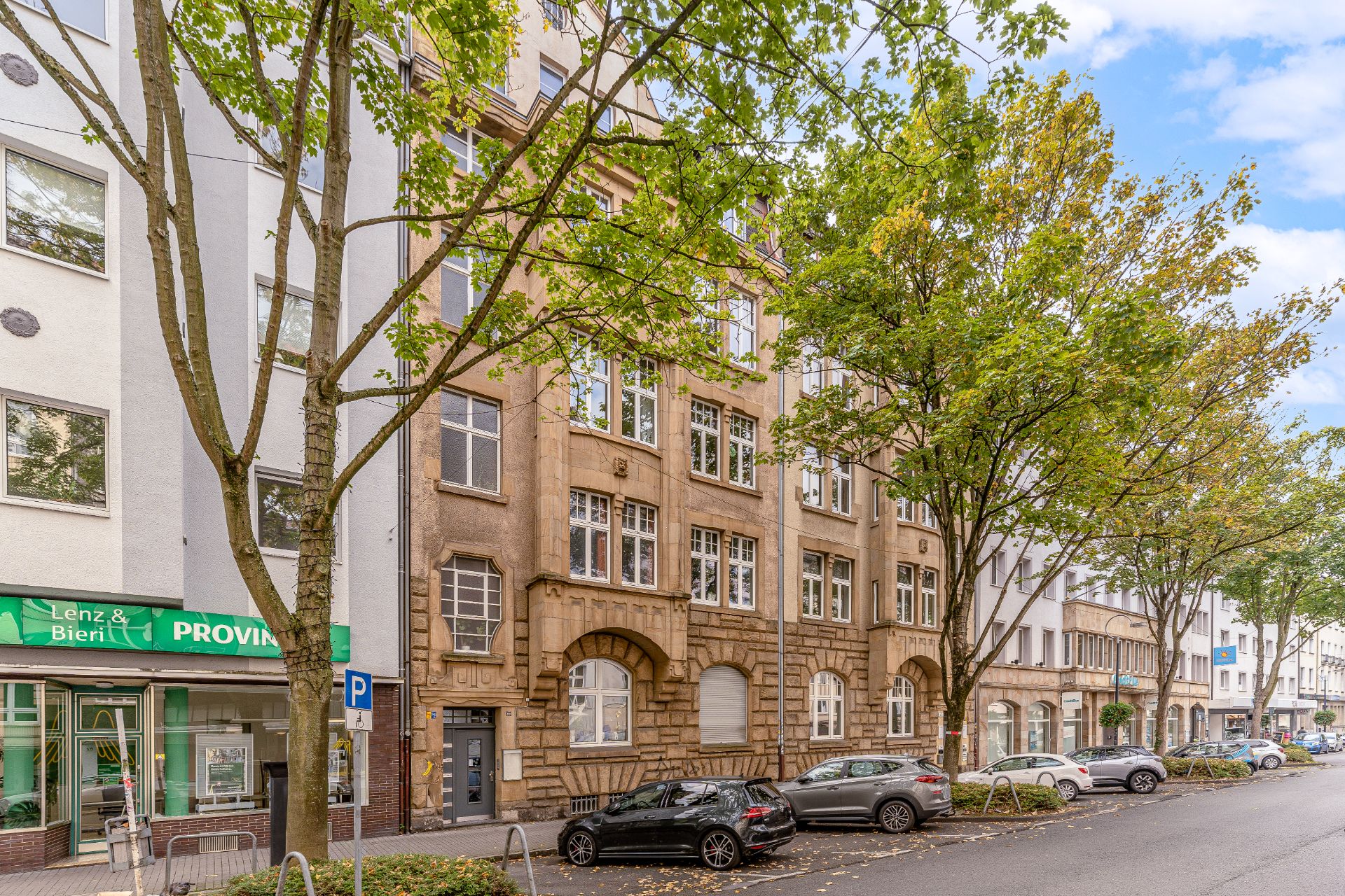 Wohnhaus mit Sandsteinfassade und großen Fenstern, Bäume und parkende Autos an der Straße.