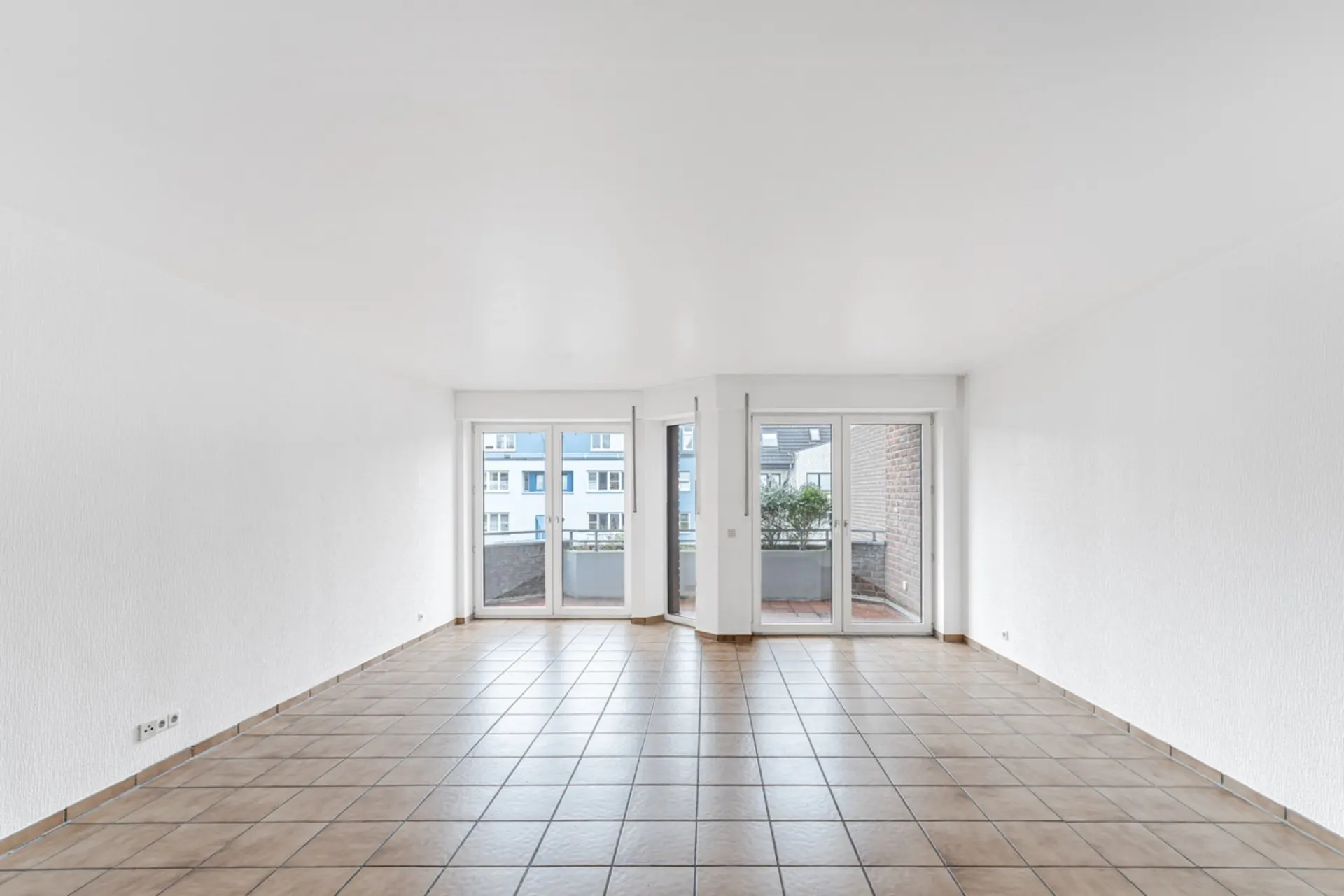 Leeres, helles Zimmer mit Fliesenboden und großen Fenstern zu einem Balkon.