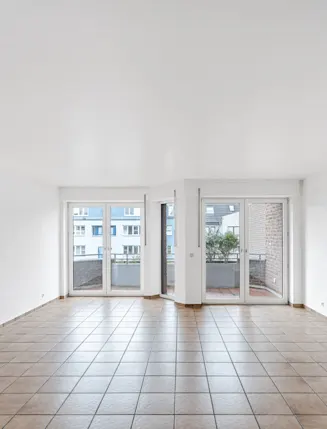 Leeres, helles Zimmer mit Fliesenboden und großen Fenstern zu einem Balkon.