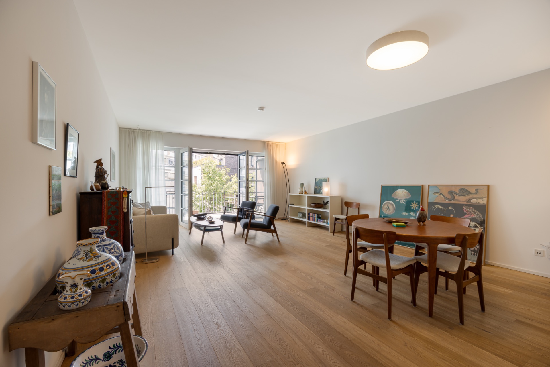 Helles Wohnzimmer mit Holzboden, Essbereich, Sofa, Sesseln und großen Fenstern zum Balkon.