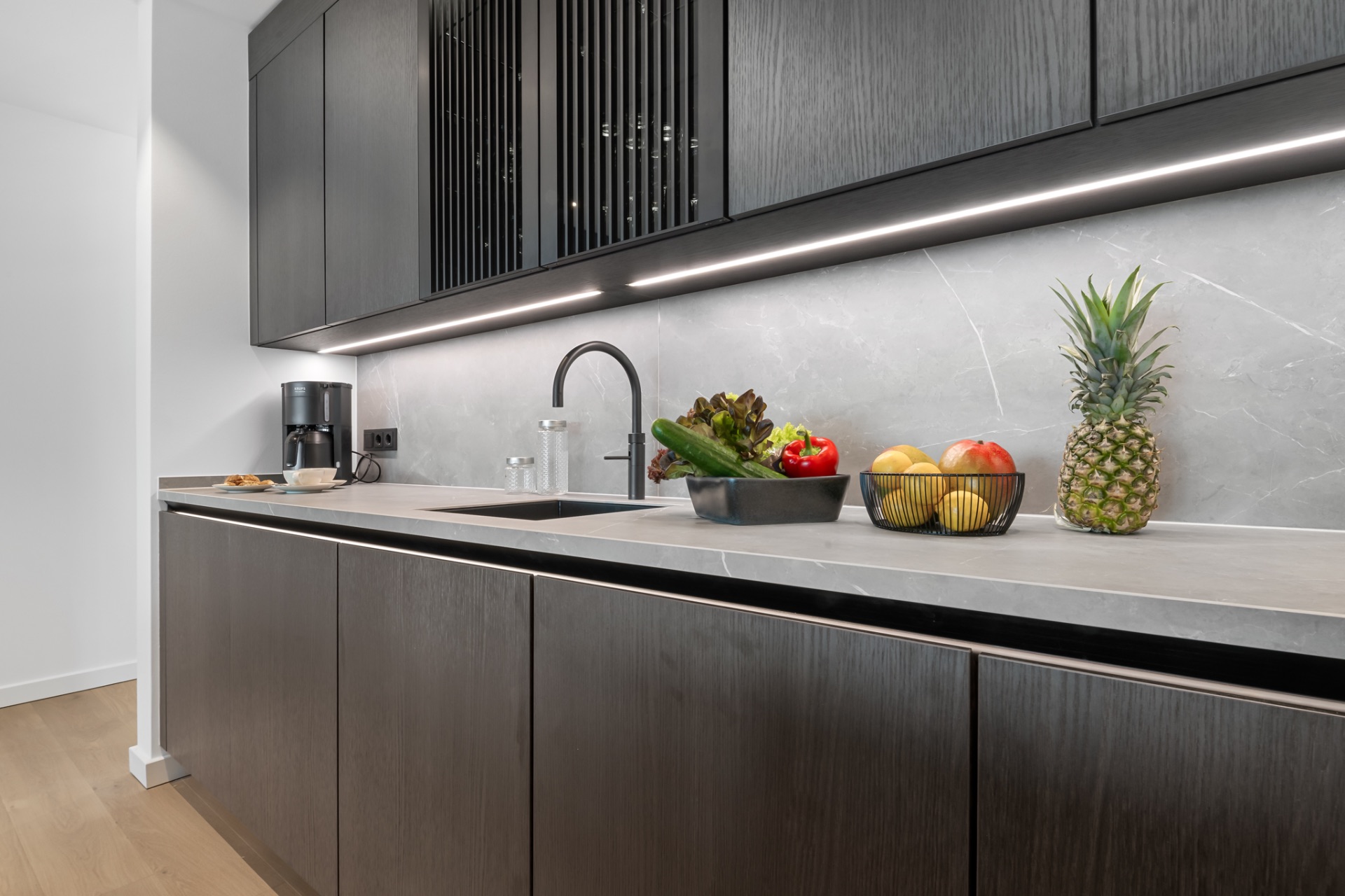 Moderne Küche mit dunklen Schränken, Obstschale, Gemüse und einer Ananas auf der Arbeitsplatte.
