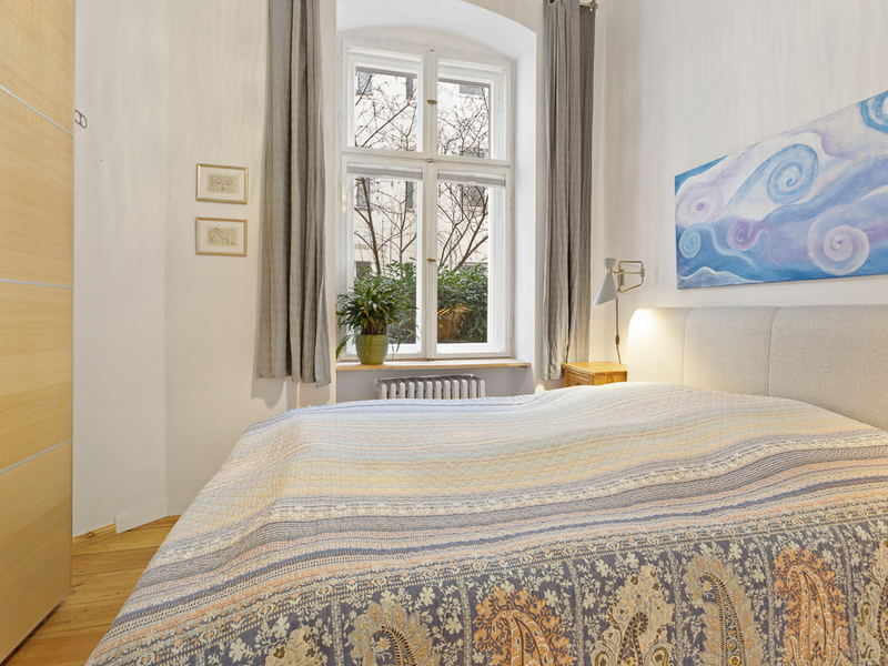 Helles Schlafzimmer mit großem Bett, Fenster, Pflanze und buntem Bild an der Wand.