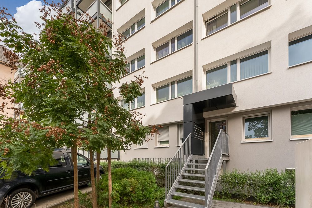 Eingang eines Mehrfamilienhauses mit Treppe, Bäumen und geparktem Auto davor.
