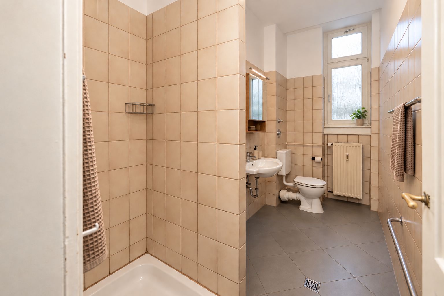 Gefliestes Badezimmer mit Dusche, Waschbecken, Spiegel, Toilette und Fenster.