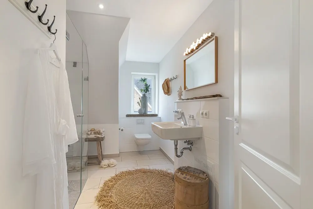 Helles Bad mit Dusche, Waschbecken, Spiegel, WC, Fenster und rundem Teppich.