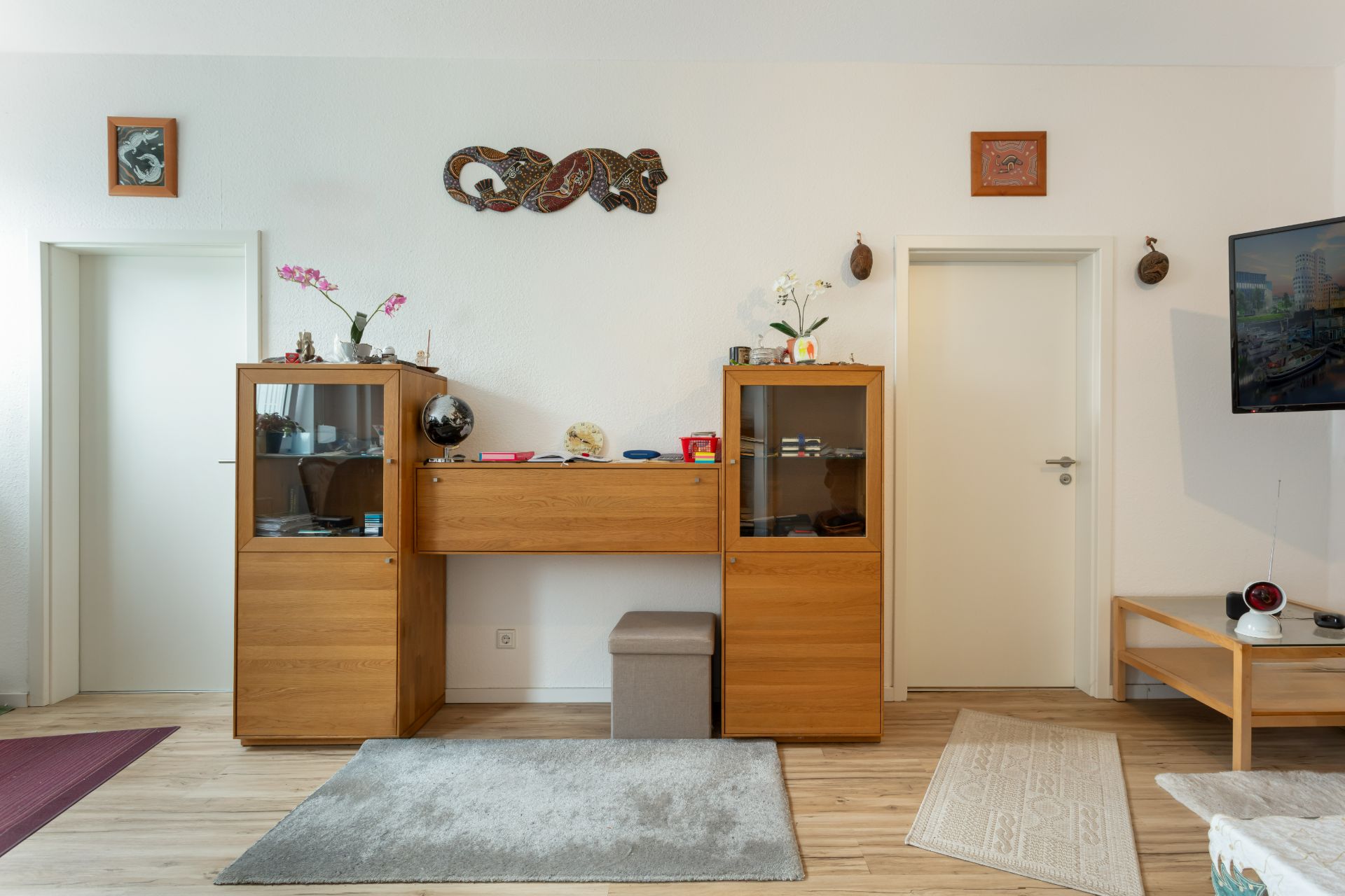 Wohnzimmer mit Holzschrank, Vitrine, Teppichen und Wanddeko zwischen zwei weißen Türen.